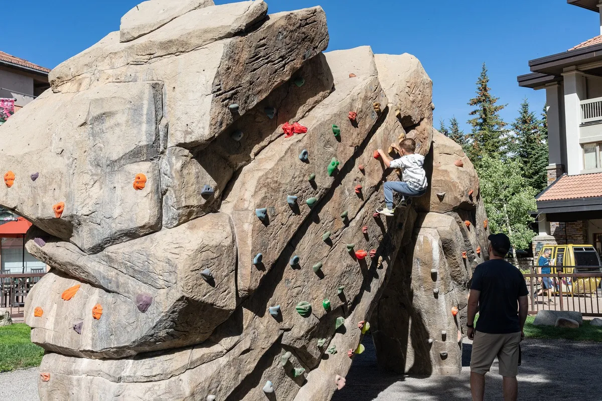 The image features a child climbing a rock wall under the supervision of an adult in a bright, outdoor setting. The playful scene emphasizes active family fun and safe environments for young children.