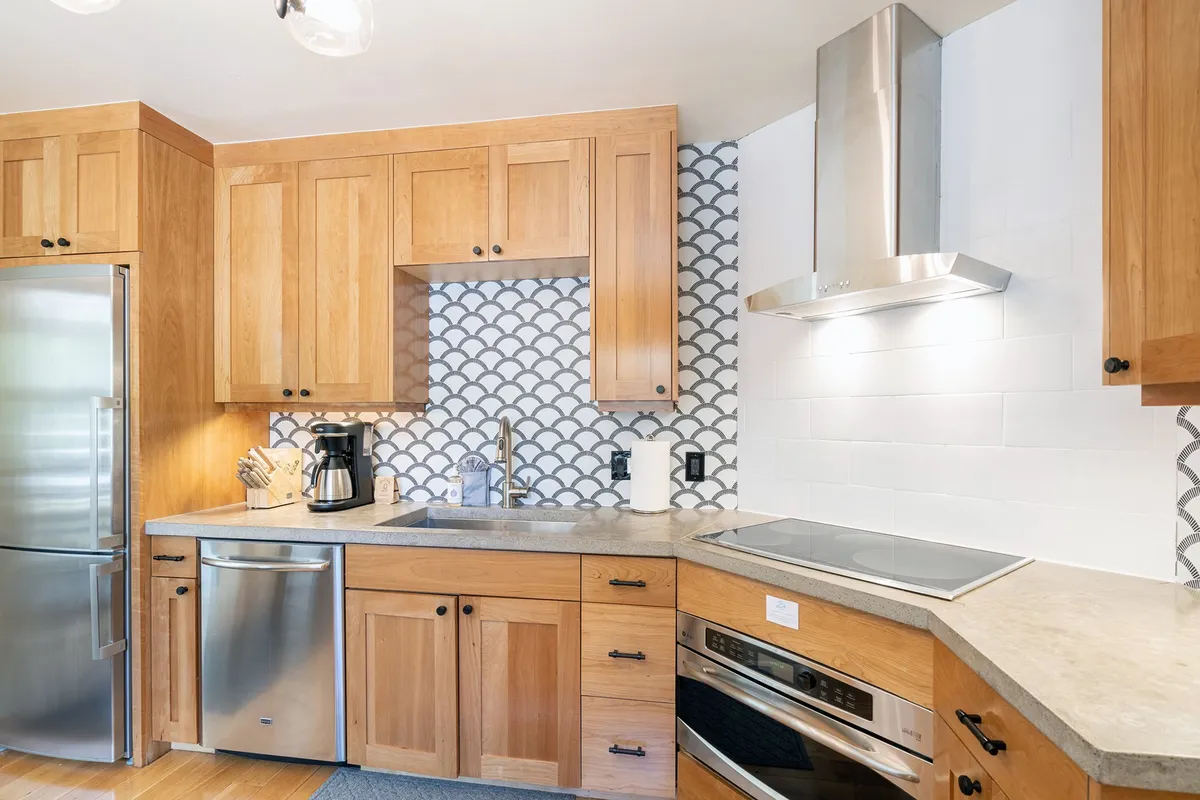 This interior image features a bright, modern kitchen with wood cabinetry, a striking patterned backsplash, stainless steel appliances, and a spacious countertop. The layout is open and inviting, perfect for preparing meals or entertaining guests.