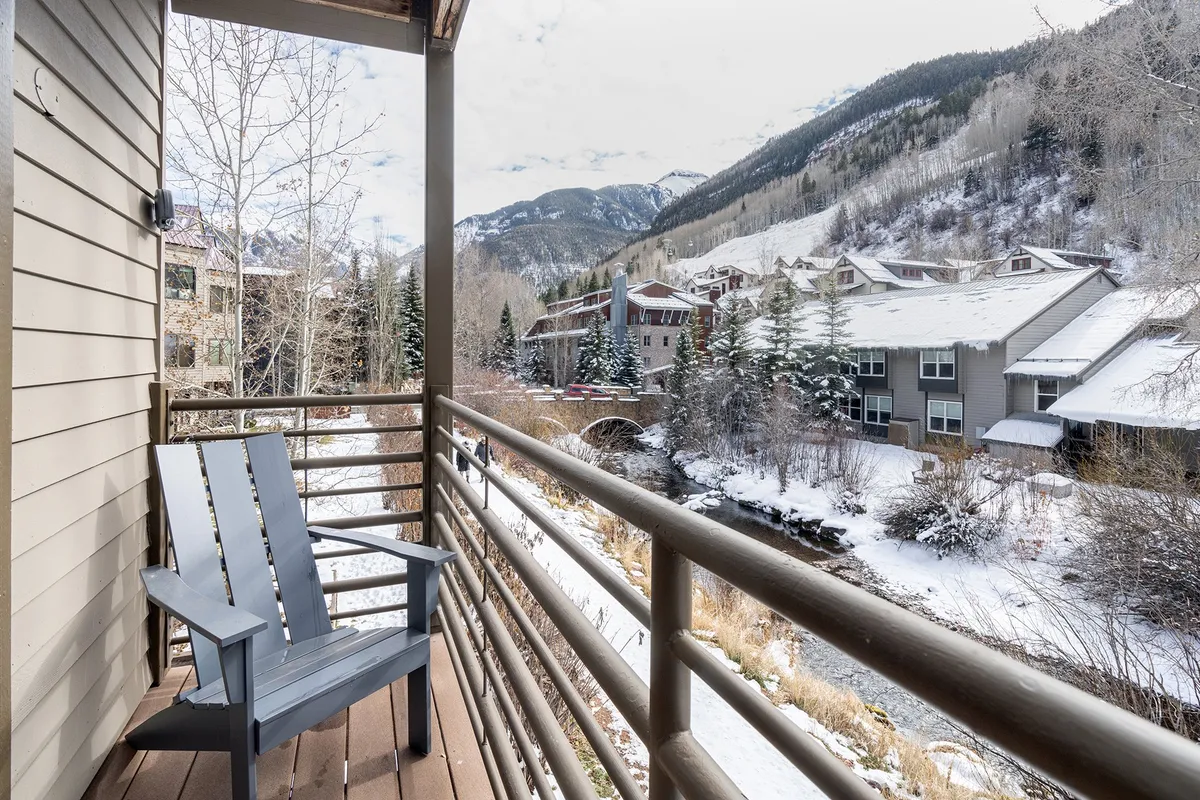 A serene winter balcony scene featuring a wooden railing, a gray chair positioned for relaxing, snow-covered mountain peaks in the distance, a gentle stream flowing by, and trees framing the view. Soft natural light pours onto the balcony, emphasizing the tranquil outdoor atmosphere and the proximity to both water and mountains.