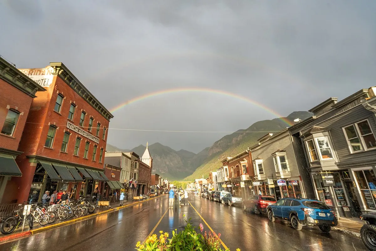 This uplifting scene depicts historic buildings, a rainbow arcing across the sky, reflections on a wet street, and bicycles parked nearby. It suggests a vibrant, walkable neighborhood with opportunities for shopping, exploration, and family outings.