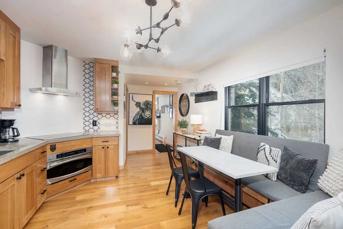 An inviting and spacious interior featuring a modern kitchen island, a dining table with chairs, colorful wall art, and notably, large windows that flood the room with natural light. The windows offer glimpses of the outside environment, making the space feel bright, open, and connected to nature.