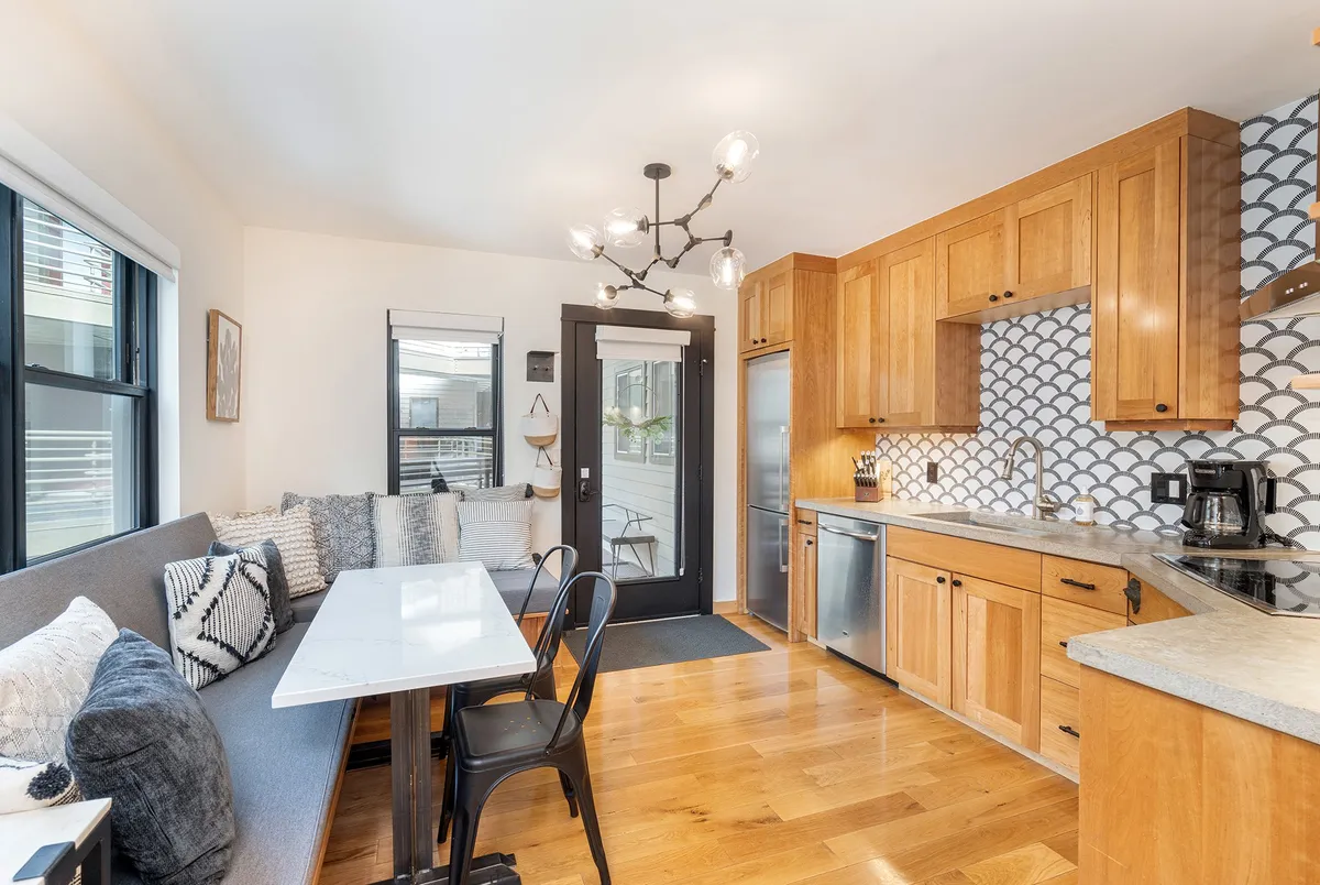 This image presents a cozy, contemporary living area with a dining table, sofa seating, and a view into the kitchen cabinetry. The lighting fixture and wall tile pattern contribute to the modern ambiance, while the arrangement of furnishings invites relaxation and social gatherings.