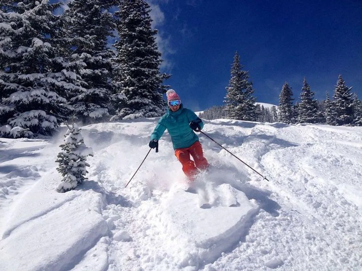 A skier glides down a pristine, snow-covered slope lined by evergreen trees, with dramatic mountains rising in the background. The scene is alive with winter energy, capturing both the thrill of skiing and the camaraderie of outdoor adventure.