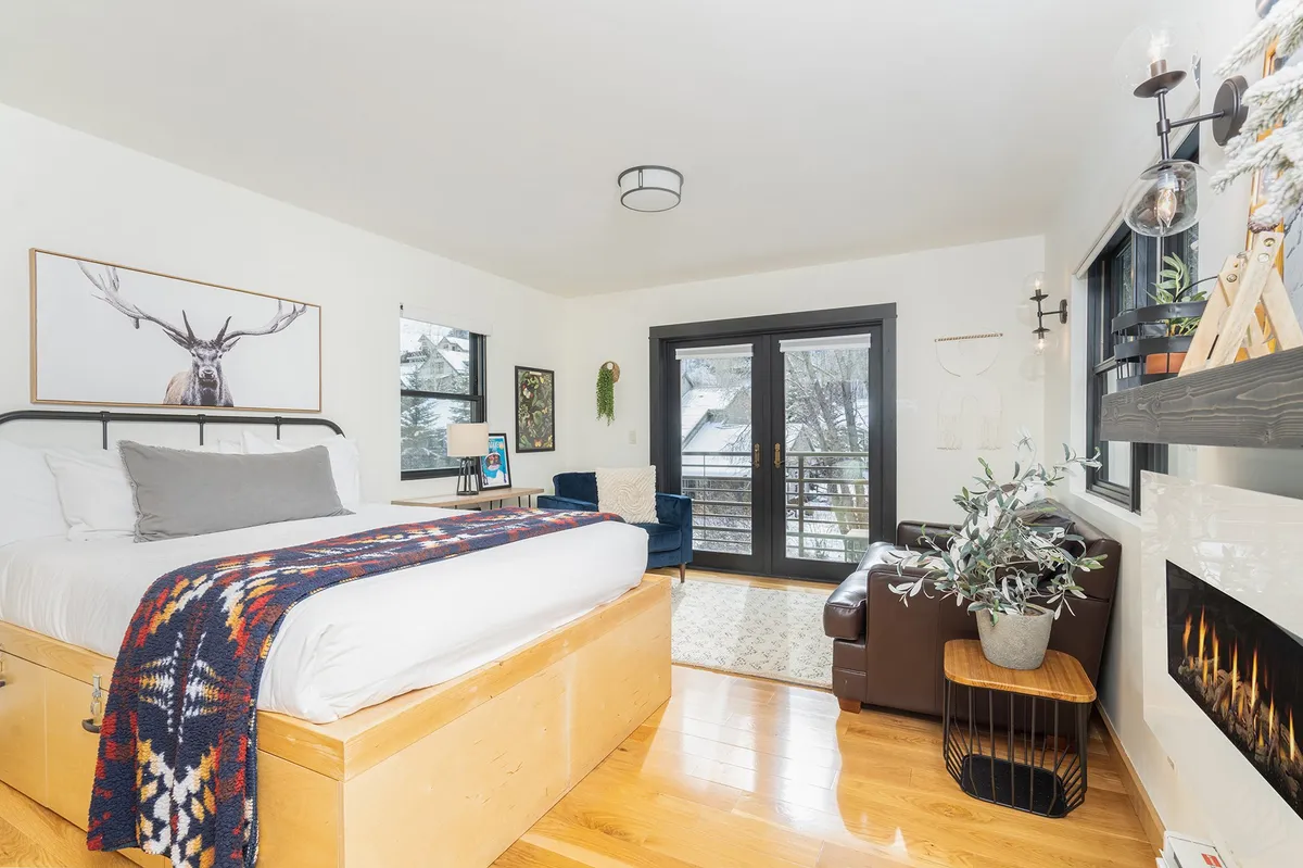 The image showcases a stylish condo interior with a cozy bed draped in a decorative blanket, large windows revealing beautiful natural light and likely scenic Telluride views, a comfortable seating area perfect for relaxing or reading, and a modern fireplace that adds warmth and ambiance.