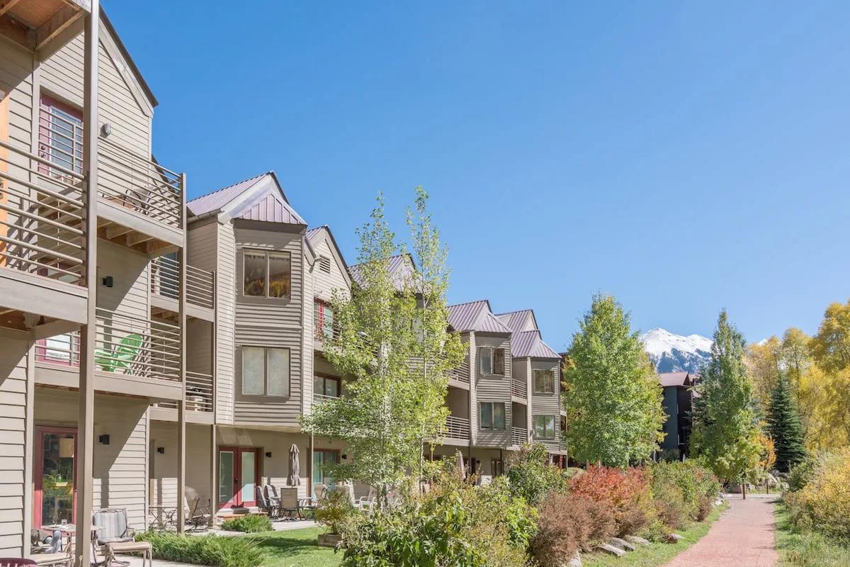 This view captures townhouse facades with balconies overlooking a landscaped pathway. In the background, snow-capped mountains rise above, and the light suggests either dawn or dusk, adding to the romantic ambiance.