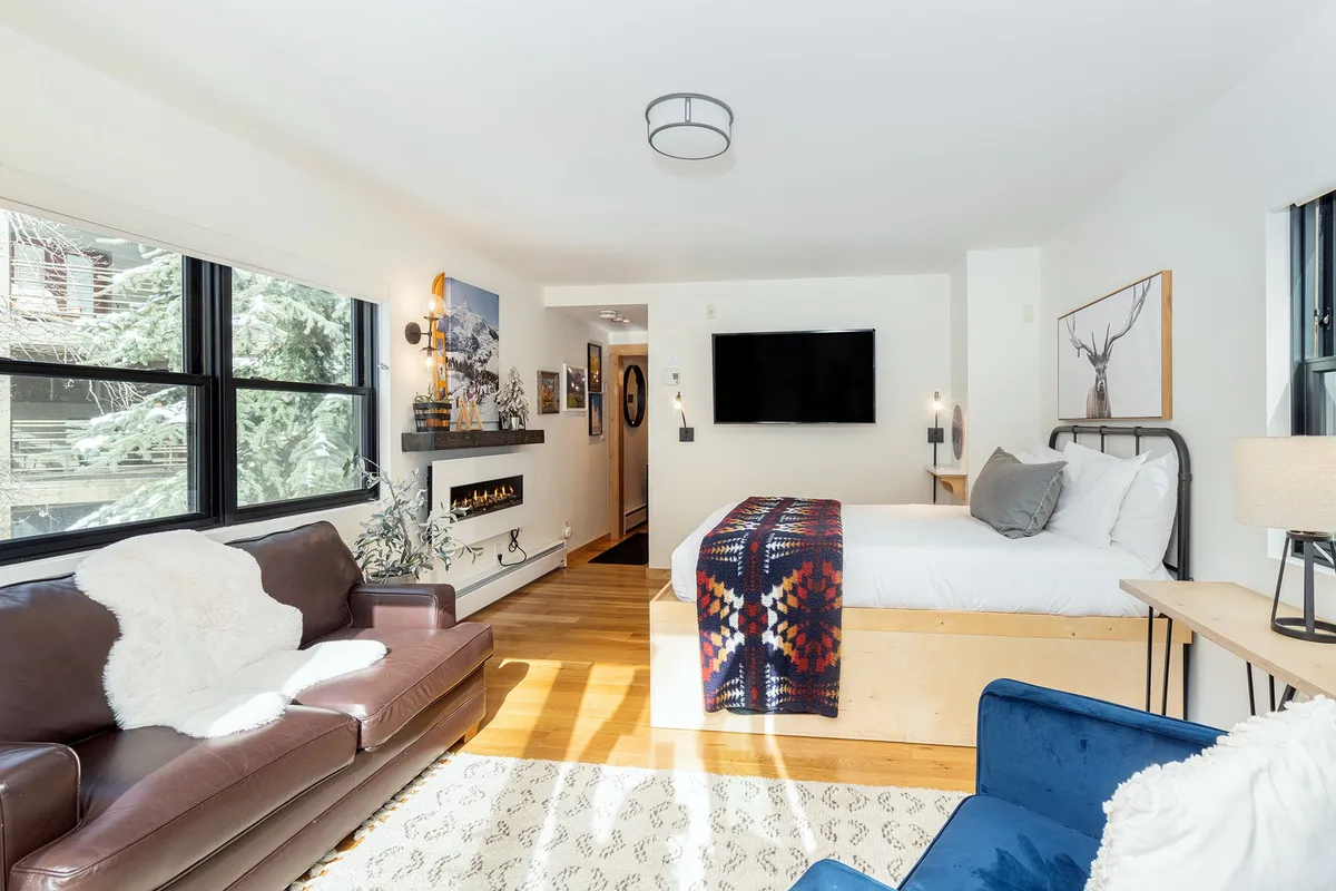 A cozy, warmly lit bedroom featuring a queen bed adorned with a colorful blanket, a brown leather sofa at the foot, a modern electric fireplace, and a blue armchair in the corner. Large windows allow natural light to fill the space, creating an inviting and relaxing ambiance perfect for unwinding.
